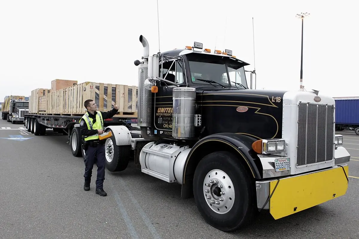 An officer in a safety vest signals a flatbed truck loaded with wooden crates through an inspection station in Seattle, Washington.