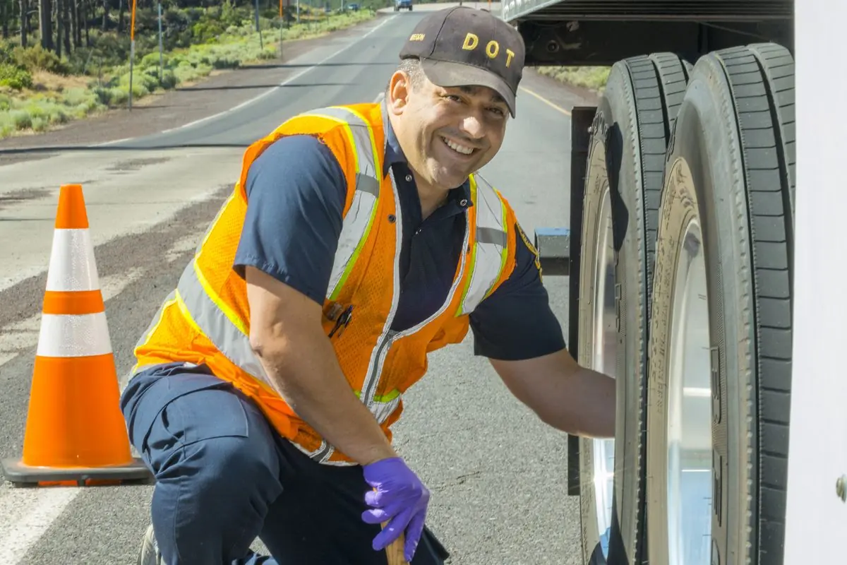 A safety specialist in an orange vest kneels beside a semi trailer and checks its tires at an Oregon inspection station.