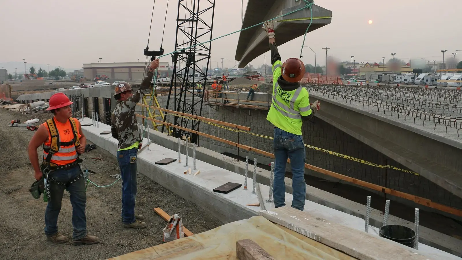 Three construction workers in hard hats guide a concrete bridge beam lifted by a crane at an Oregon highway interchange.