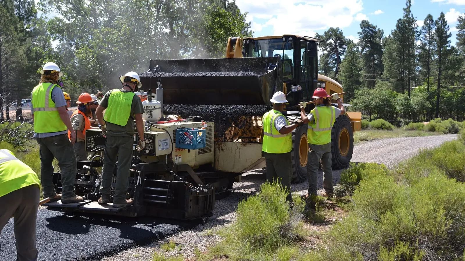 A crew in hard hats and safety vests works beside a paving machine as a front loader refills it with asphalt in Arizona.