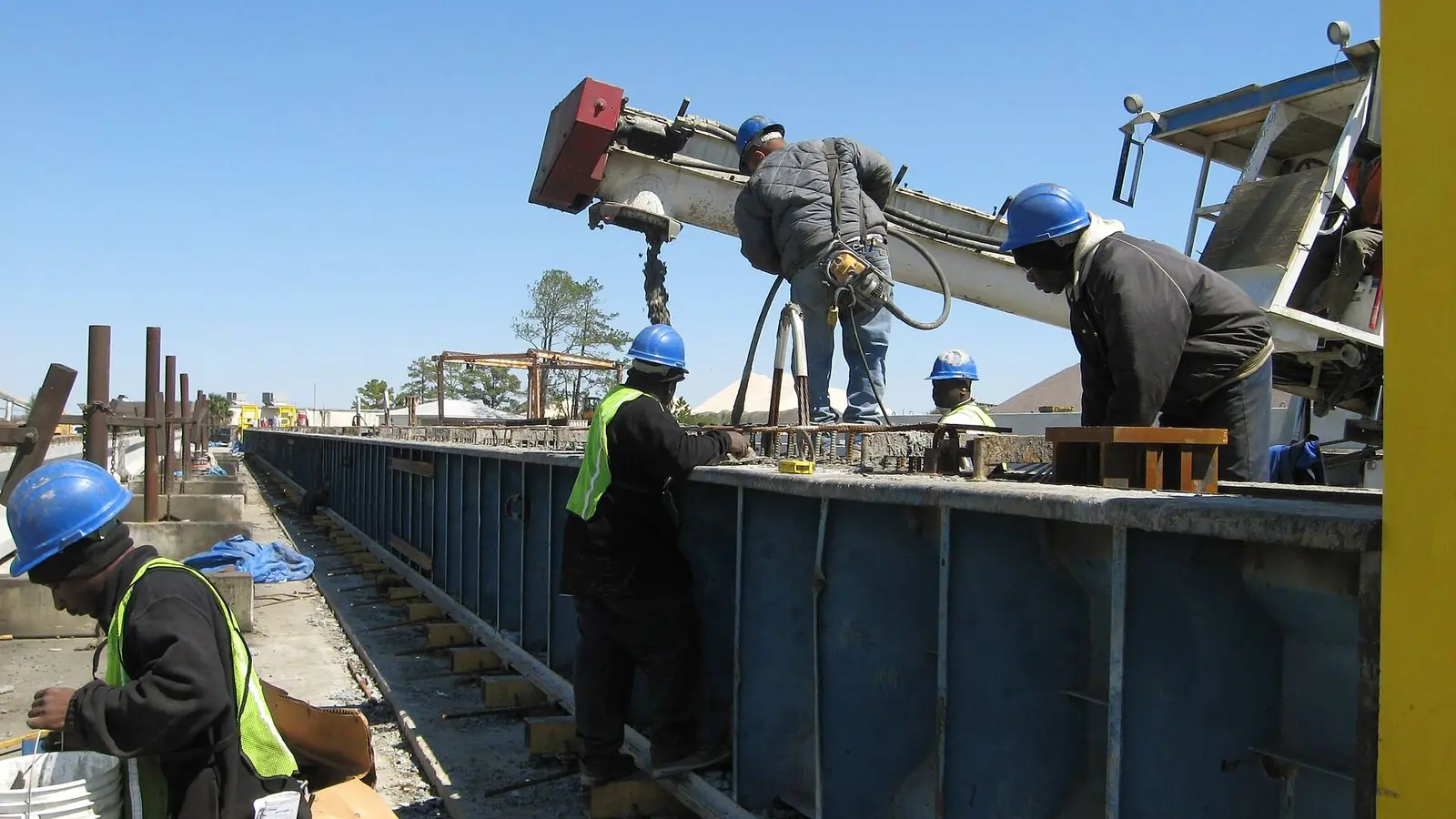 Workers in blue hard hats place concrete into a long steel form for bridge beams at a precast casting plant in Savannah, Georgia.