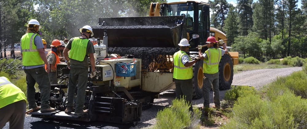 A crew in hard hats and safety vests works beside a paving machine as a front loader refills it with asphalt.